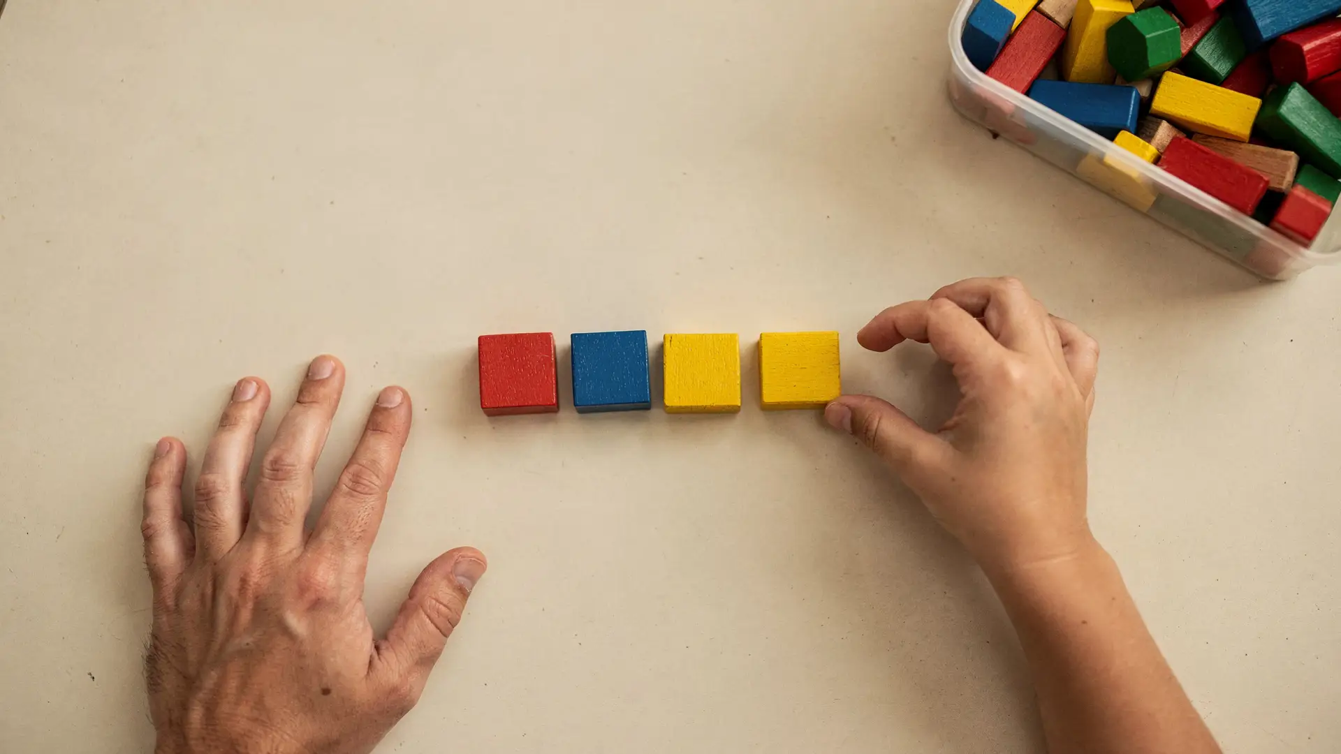 Mão de adulto ao lado de uma fila de blocos vermelho, azul e amarelo que outra mão completa, jogo de sequência em atividades para crianças com TDAH