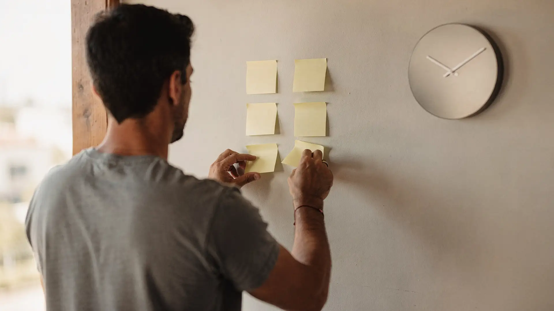 Homem de costas cola post-its em branco numa parede ao lado de um relógio, dividindo tarefas em partes menores, estratégia útil no TDAH em adultos
